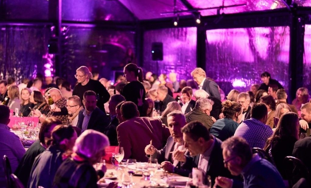 Long dinner tables, crowds enjoying fare within a purple-lit pop-up marquee