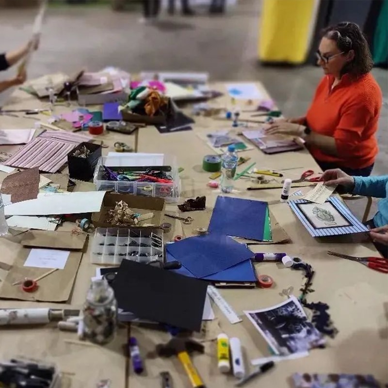 A crafts workshop at a market with heaps of paper, paints and bits and bobs covering a table