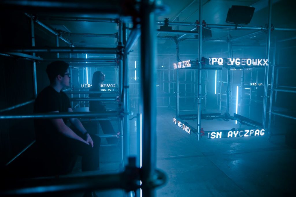 Two people sitting in darkened room looking at cage-like light installation