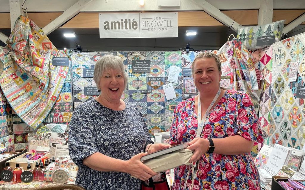 Two people holding books in front of a quilting market stall