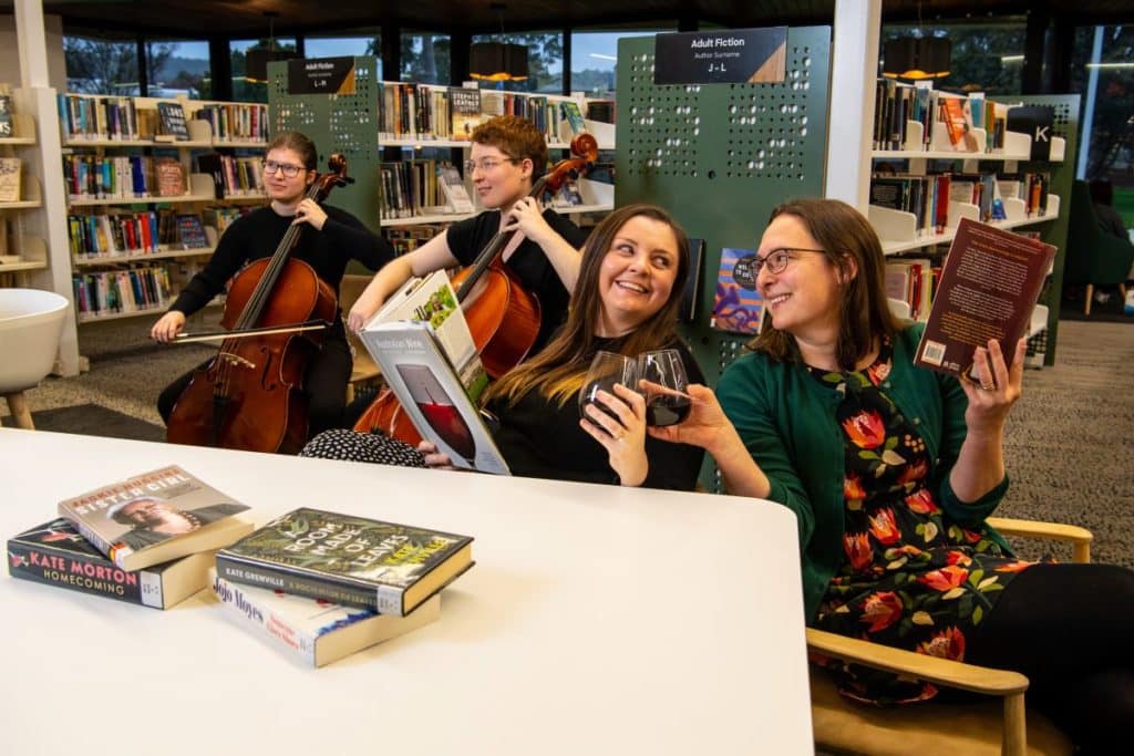 Four people having a laugh in a library - two people playing the cello, two people reading books