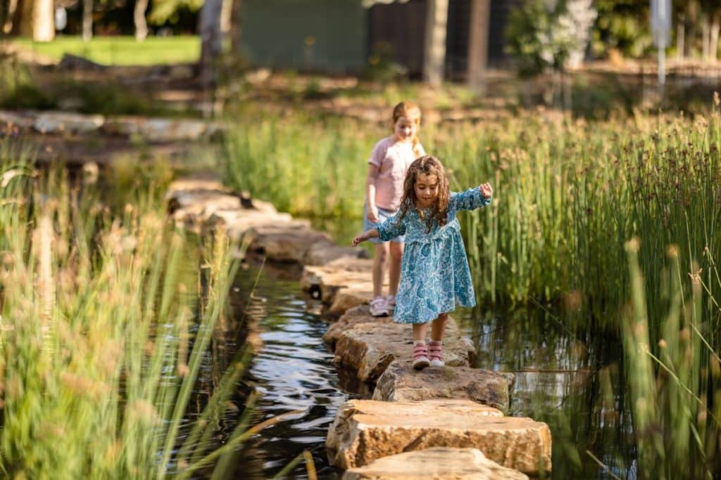 Two kids following a path over a stream surrounded by tall water grass